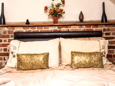 A bed with cream and gold pillows against a brick wall with decorative items on a wooden shelf at Woodcock in Wheatacre