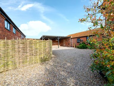 An outdoor gravel area with a woven fence carport and brick buildings with windows and plants at Woodcock in Wheatacre