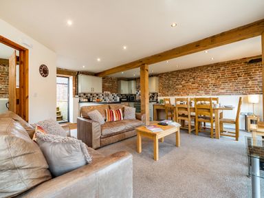 An open living area with two brown leather sofas wooden coffee and dining tables exposed brick walls and kitchen cabinets at Snipe in Wheatacre