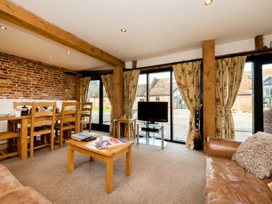 A living room with wooden dining table chairs a coffee table a television on a stand and large windows with floral curtains at Snipe in Wheatacre