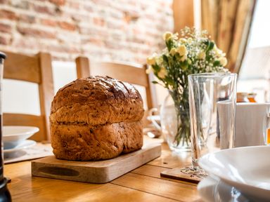 A loaf of bread on a wooden cutting board with a glass and a vase of flowers on a dining table at Snipe in Wheatacre