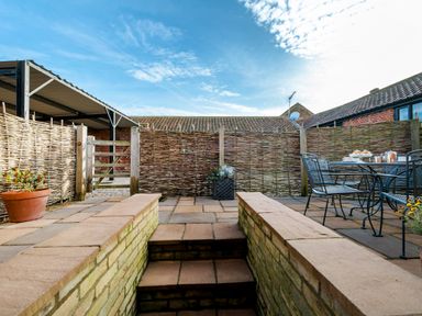 An outdoor patio area with stone steps leading up to a paved space with metal chairs and a table a potted plant and woven fencing at Snipe in Wheatacre