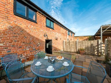 An outdoor patio with a round metal table and chairs set with cups and scones next to a brick building with black windows and a woven fence at Snipe in Wheatacre