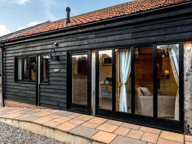 A black wooden exterior wall with windows and a glass door showing a living area at Pheasant in Wheatacre