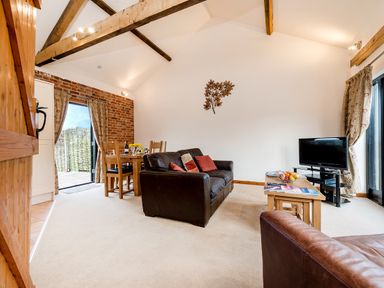 A living room with a wooden dining table and chairs a brown leather sofa a coffee table with fruit and magazines a television and exposed wooden beams at Pheasant in Wheatacre