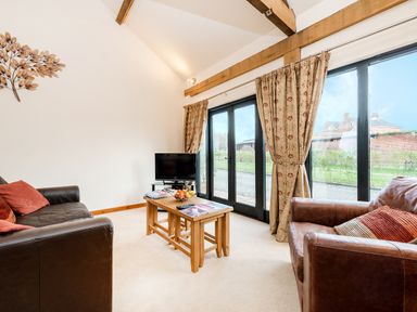 A living room with two leather armchairs a wooden coffee table a small television and large glass doors with curtains at Pheasant in Wheatacre