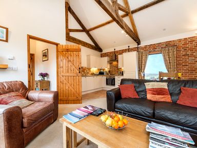 A living room with leather sofas a wooden coffee table with fruit and magazines an open door leading to a kitchen with white cabinets and exposed wooden beams at Pheasant in Wheatacre