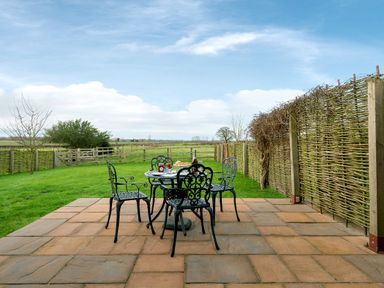 An outdoor patio with a metal table and four chairs on a tiled surface next to a woven fence and green fields at Pheasant in Wheatacre