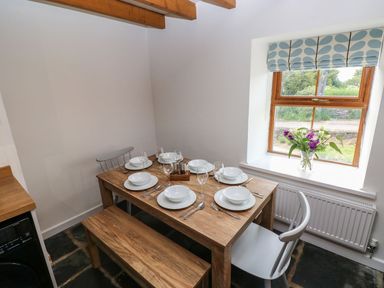 A dining area with a wooden table set for six people near a window with a vase of flowers at Dalton Cottage in Llansaint