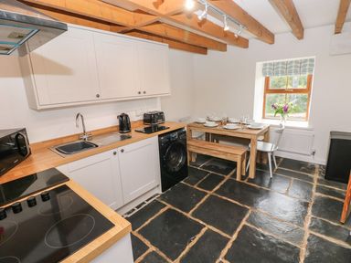 A kitchen with wooden beams a dining table with chairs and a bench and kitchen appliances at Dalton Cottage in Llansaint