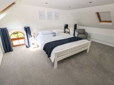 A bedroom with a white bed with pillows and blankets two bedside tables with lamps a gray armchair and two windows with dark curtains at Dalton Cottage in Llansaint