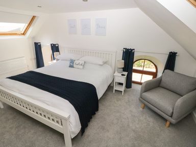 A bedroom with a white bed frame white bedding dark blue blanket two blackout curtains a grey armchair and two small white nightstands Dalton Cottage Llansaint