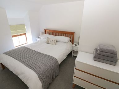 A bedroom with a wooden bed frame a white dresser with folded towels and a window with a blind at Dalton Cottage in Llansaint