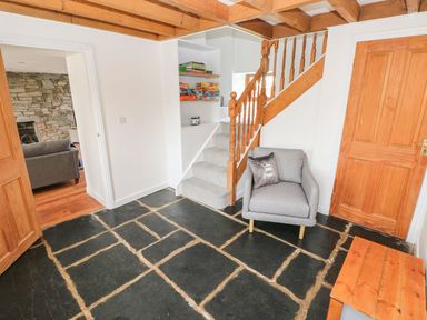 A small room with stone tile flooring a gray armchair wooden stairs and bookshelf under stairs at Dalton Cottage in Llansaint