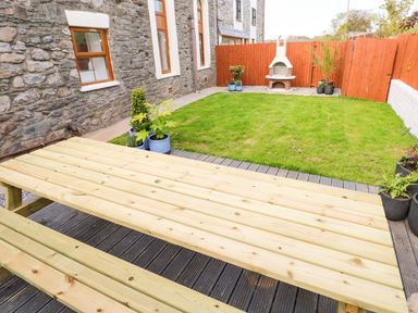 A backyard with a wooden picnic table grass area potted plants a stone wall and a barbecue grill at Dalton Cottage in Llansaint