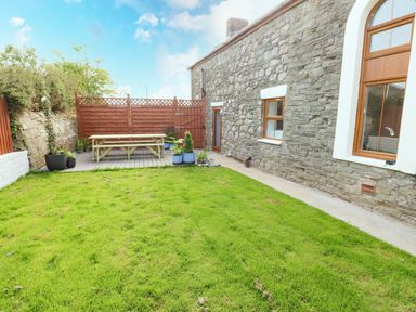 A backyard with green grass a wooden picnic table and potted plants beside a stone house with windows at Dalton Cottage in Llansaint