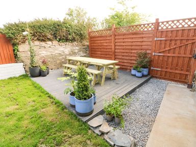 A backyard with a wooden picnic table and benches on a deck surrounded by potted plants and fenced area at Dalton Cottage in Llansaint