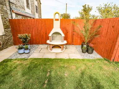 A small backyard with a stone barbecue grill and potted plants against a red wooden fence at Dalton Cottage in Llansaint