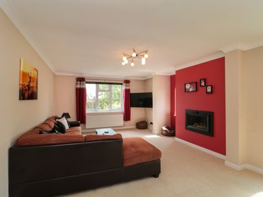 A living room with a brown sectional sofa a mounted TV red curtains a fireplace and framed pictures on a red accent wall at Te Papa in North Petherton