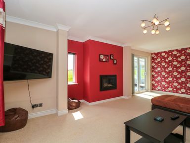 A living room with a wall-mounted TV a fireplace red accent walls floral wallpaper a brown ottoman and a black coffee table at Te Papa in North Petherton