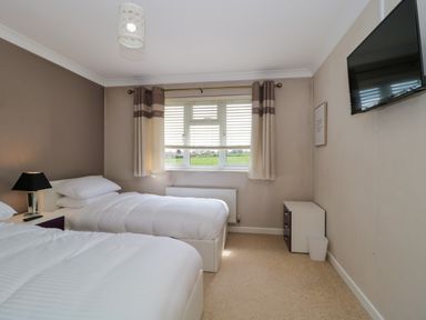 A bedroom with two single beds a window with curtains a bedside table lamp a mounted television and a small dresser with a trash can at Te Papa in North Petherton