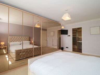 A bedroom with a large mirrored wardrobe a bed with a tufted headboard bedside tables with lamps a wall-mounted TV and an open door leading to a bathroom at Te Papa in North Petherton