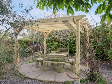 A wooden pergola over a circular paved area with wooden benches and a table in a garden at Te Papa in North Petherton
