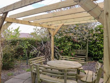 A wooden pergola with string lights above a round wooden table and benches in a garden at Te Papa in North Petherton
