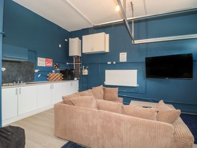 A living area with a beige sofa a wall-mounted television a radiator and a kitchenette at Te Papa in North Petherton