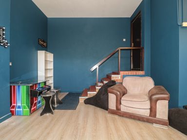 A room with blue walls a brown and beige armchair stairs a white shelf a black bean bag two stools and a colorful arcade game cabinet at Te Papa in North Petherton