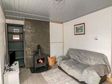 A living room with a sofa covered by a grey cover a wood stove with fire a tv on a white cabinet and a bookshelf at Curragh Cottage in Rathimney near New Ross County Wexford