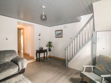 A living room with a sofa covered with a grey quilted cover a floral armchair a small table with a radio and a plant a framed picture on the wall and a staircase at Curragh Cottage Rathimney near New Ross County Wexford