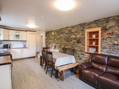 A kitchen and dining area with wooden chairs, a table covered with a cloth, a wooden bench, a brown leather sofa, and stone walls at Curragh Cottage in Rathimney near New Ross County Wexford