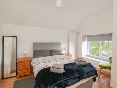 A bedroom with a double bed grey headboard two wooden bedside tables with lamps a full length mirror a window with grey blind and a green chair at Curragh Cottage Rathimney near New Ross County Wexford