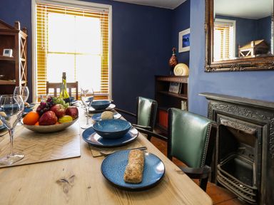 A dining room with a wooden table set with blue plates glasses and a fruit bowl near a window and a fireplace at Georgian Hideaway in Weymouth