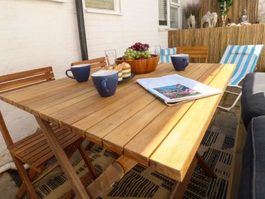An outdoor wooden table with three blue mugs a bowl of fruit a plate of muffins and an open magazine at Georgian Hideaway in Weymouth