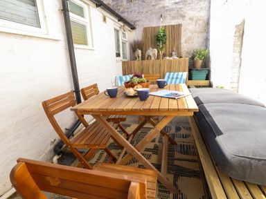 A wooden table with wooden chairs and cushioned bench in a small outdoor patio with fruit and mugs on the table at Georgian Hideaway in Weymouth