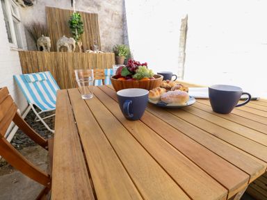 An outdoor wooden table with blue mugs plates of pastries and a bowl of fruit with striped chairs in a courtyard at Georgian Hideaway Weymouth