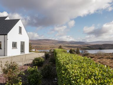 A white house with a dark roof beside a gravel path and green hedge with hills and a lake in the background at Moorhen House in Loughanure near Annagry County Donegal