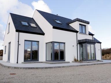 A modern white house with large gray framed windows and a gravel driveway at Moorhen House in Loughanure near Annagry County Donegal