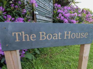 A wooden sign with the text The Boat House on a lawn with purple flowers and a green shed in the background at The Boathouse in Belford
