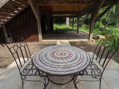 A round mosaic table with two metal chairs on a stone patio under a wooden deck at The Boathouse in Belford
