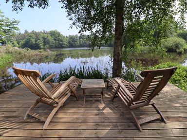 A wooden deck with two wooden lounge chairs and a table overlooking a lake with trees at The Boathouse in Belford