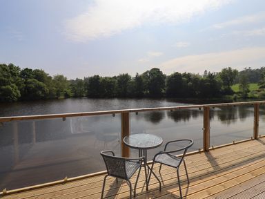 A wooden deck with a glass round table and two chairs overlooking a lake with trees at Otters Cove in Belford