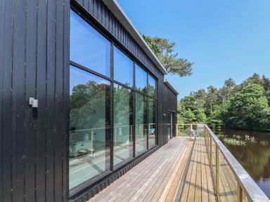 A wooden deck with glass railing outside a black building overlooking a river and trees at Otters Cove in Belford