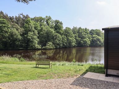 A bench on grass facing a lake with trees across the water and part of a building visible at Otters Cove in Belford