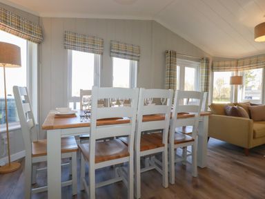 A dining area with a wooden table and white chairs next to windows with plaid curtains at 4 Meadow Retreat in Dobwalls