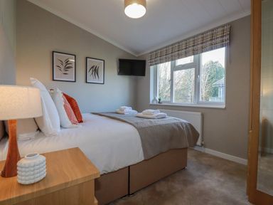 A bedroom with a bed towels on a duvet a nightstand a lamp framed pictures a wall mounted television and a window at 4 Meadow Retreat in Dobwalls