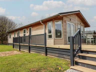 A wooden cabin with a deck and black railing in a grassy area at 4 Meadow Retreat in Dobwalls