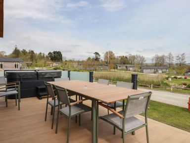 A wooden outdoor deck with a dining table and chairs and a hot tub overlooking grass and cabins at 4 Meadow Retreat in Dobwalls